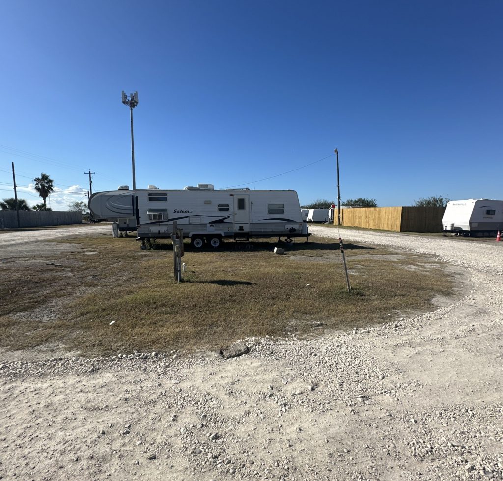 A white travel trailer is parked on a gravel and grass lot within an RV park. A tall cell tower and several palm trees are visible in the background, along with other RVs lined up near a wooden fence. The sky is clear and bright blue above the open space.