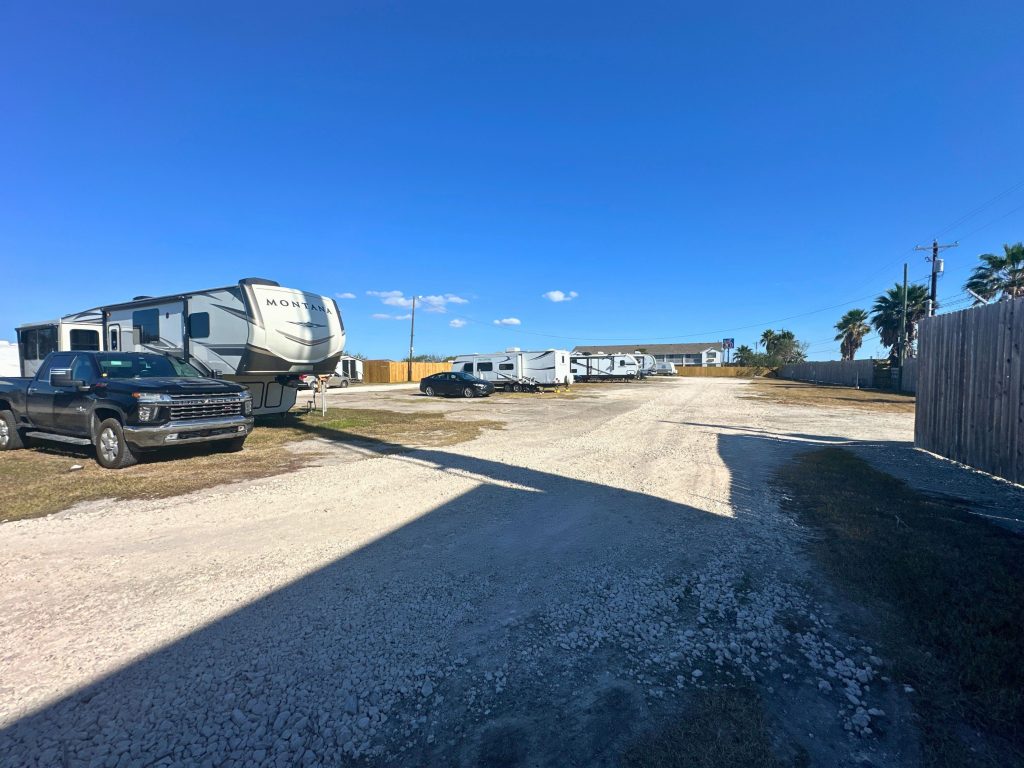 A gravel RV park lot with several RVs and vehicles parked along the sides. A large fifth-wheel RV and a black pickup truck are in the foreground on the left, while more RVs are lined up further back. A small building is visible in the distance, with palm trees and utility poles along the right side under a clear blue sky.