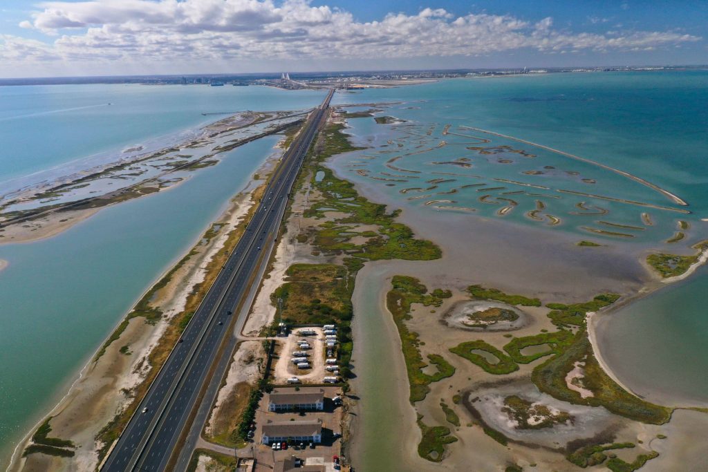 Aerial view of an RV park located along a highway and coastal wetlands. Several RVs are parked in organized rows on gravel lots, with a building at the entrance. To the left, a multi-lane highway runs parallel to a body of water, while to the right are marshlands and tidal pools with patches of green vegetation.